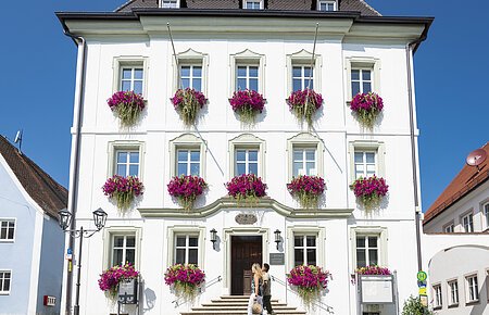 Das Rathaus in Monheim: Ein weißes Gebäude mit mehreren Fenstern, an denen Blumenkästen mit pinken Blumen hängen. Zwei Personen gehen davor vorbei.