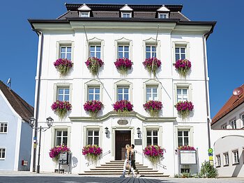 Das Rathaus in Monheim: Ein weißes Gebäude mit mehreren Fenstern, an denen Blumenkästen mit pinken Blumen hängen. Zwei Personen gehen davor vorbei.