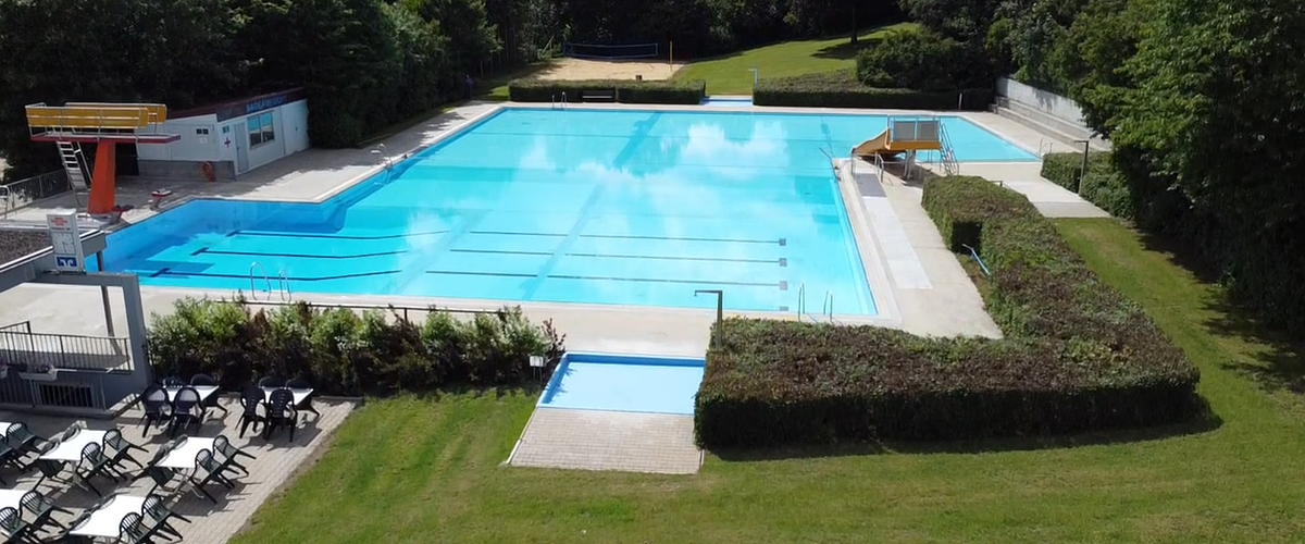 Außenansicht eines leeren Schwimmbeckens mit Sprungturm, Rutsche, Sitzbereich und umgebender Grünfläche.