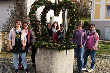 Gruppe von acht Personen steht um einen mit Blumen und Zweigen geschmückten Brunnen auf einem gepflasterten Platz.