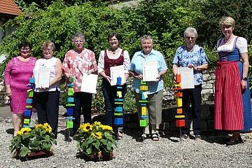 Sieben Personen stehen draußen in einer Reihe, halten Urkunden und bunte Stäbe, zwei Sonnenblumenarrangements im Vordergrund.