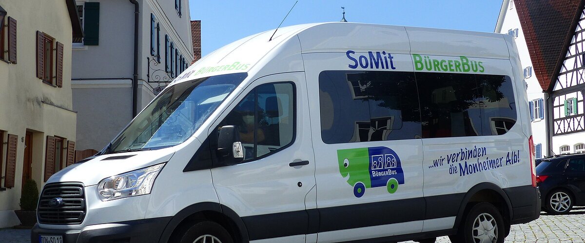 Weißer Kleinbus steht auf einem gepflasterten Platz, in der Monheimer Innenstadt . Auf dem Bus ist die Aufschrift "SoMit BürgerBus" und "wir verbinden die Monheimer Alb".