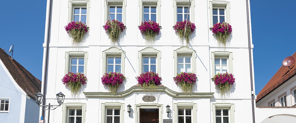 Das Rathaus in Monheim: Ein weißes dreistöckiges Gebäude mit mehreren Fenstern, an denen Blumenkästen mit pinken Blumen hängen. Zwei Personen gehen davor vorbei.