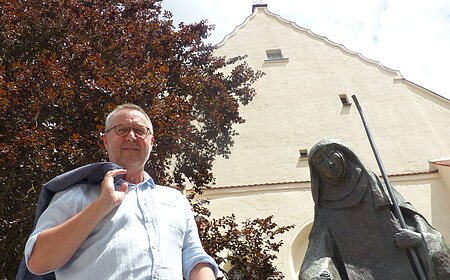 Mann mit Jacke über der Schulter steht neben einer Statue, der Walburga-Statue in Monheim vor einem gelbgestrichenem Gebäude mit Giebeldach. Im Hintergrund ein großer Baum und sonniger Himmel.