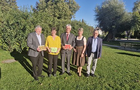 Fünf Personen stehen auf einer Wiese vor Bäumen, drei halten bunte Geschenke in den Händen, im Hintergrund blauer Himmel. Renteneintritt der drei langjährigen Mitarbeiter der VG Monheim.