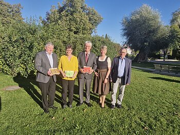 Fünf Personen stehen auf einer Wiese vor Bäumen, drei halten bunte Geschenke in den Händen, im Hintergrund blauer Himmel. Renteneintritt der drei langjährigen Mitarbeiter der VG Monheim.