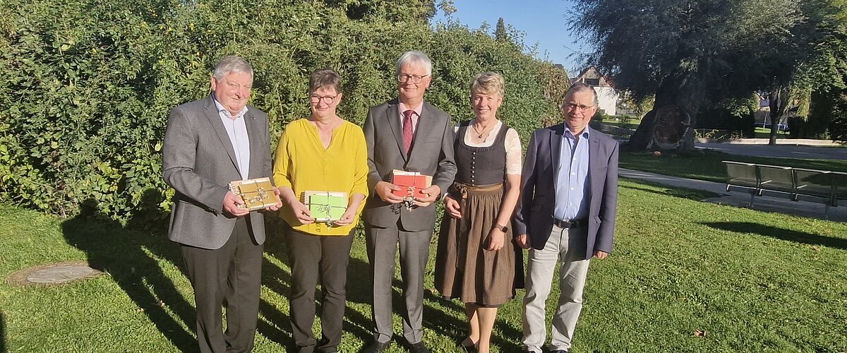 Fünf Personen stehen auf einer Wiese vor Bäumen, drei halten bunte Geschenke in den Händen, im Hintergrund blauer Himmel. Renteneintritt der drei langjährigen Mitarbeiter der VG Monheim.