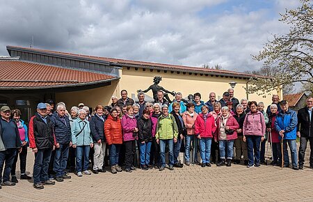 Große Gruppe von Menschen steht vor einem gelben Gebäude mit rotem Dach und einer Statue hinter Ihnen. Im Hintergrund ein Baum auf einer Wiese und bewölkter Himmel.