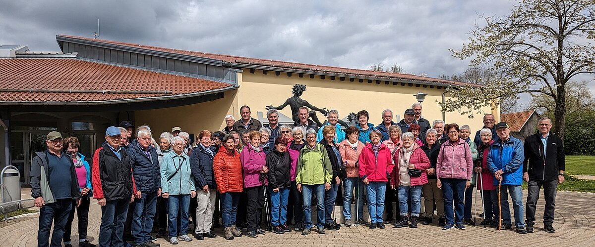 Große Gruppe von Menschen steht vor einem gelben Gebäude mit rotem Dach und einer Statue hinter Ihnen. Im Hintergrund ein Baum auf einer Wiese und bewölkter Himmel.