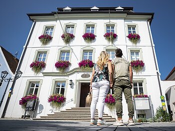 Paar, Mann und Frau von hinten vor einem weißem Gebäude mit Blumen in Blumenkästen an den Fenstern, schauen auf das Gebäude bei klarem Himmel. Eine Infovitrine auf der rechten Seite am Gebäude.