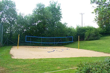 Sandvolleyballfeld mit blauem Netz und grüner Umgebung, keine Personen sichtbar.