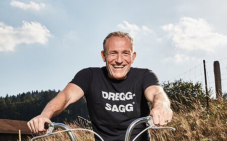 Mann mit schwarzem T-Shirt mit der weißen Aufschrift "DREGG-SAGG" und Jeans sitzt lachend auf orangefarbenem Fahrrad auf Feldweg bei Sonnenschein