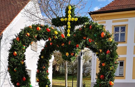 Brunnen mit grüner Osterkrone und bunten Ostereiern in einem Dorf vor Häusern bei sonnigem Wetter.
