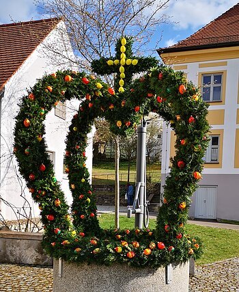 Brunnen mit grüner Osterkrone und bunten Ostereiern in einem Dorf vor Häusern bei sonnigem Wetter.