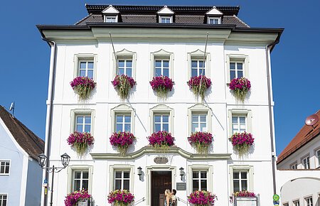 Das Rathaus in Monheim: Ein weißes dreistöckiges Gebäude mit mehreren Fenstern, an denen Blumenkästen mit pinken Blumen hängen. Zwei Personen gehen davor vorbei. Links und rechts davon jeweils ein weiteres Haus.