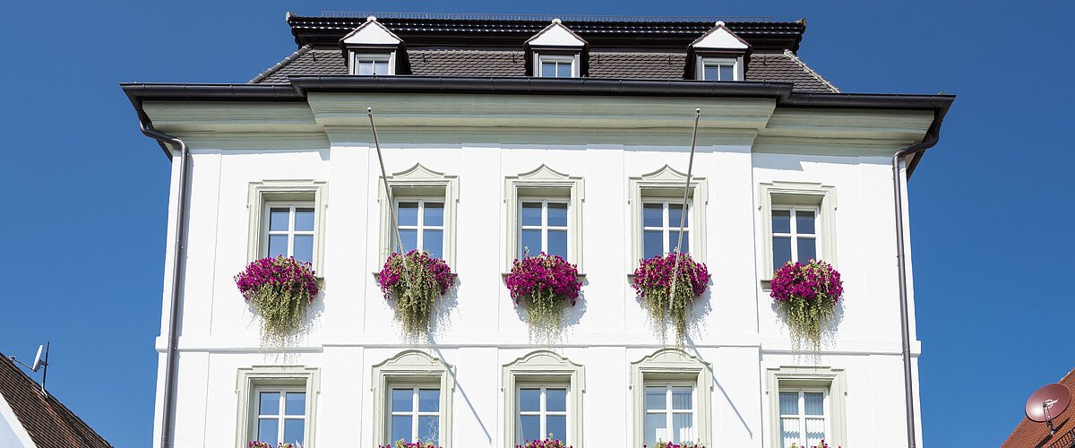 Das Rathaus in Monheim: Ein weißes dreistöckiges Gebäude mit mehreren Fenstern, an denen Blumenkästen mit pinken Blumen hängen. Zwei Personen gehen davor vorbei. Links und rechts davon jeweils ein weiteres Haus.