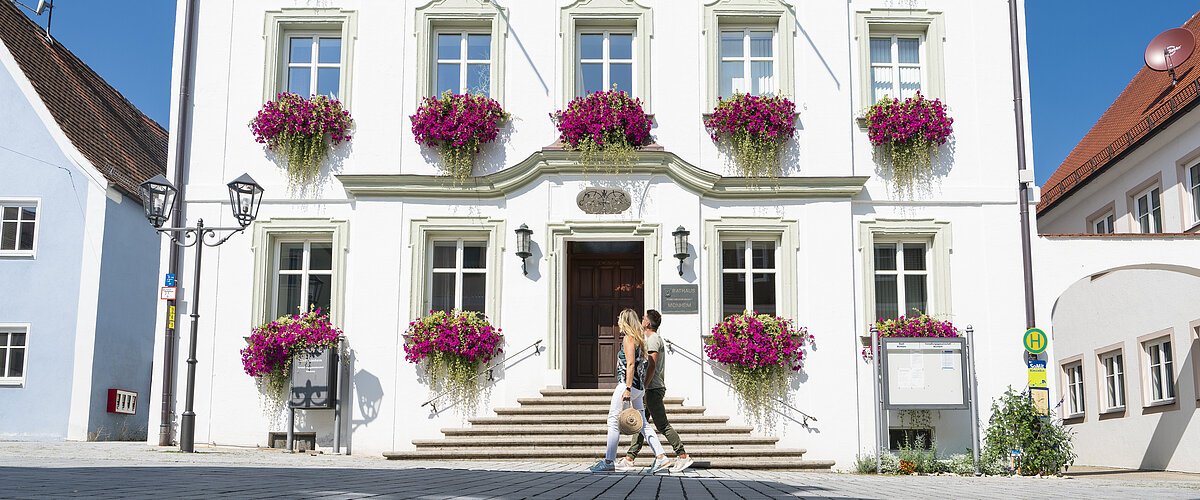 Das Rathaus in Monheim: Ein weißes dreistöckiges Gebäude mit mehreren Fenstern, an denen Blumenkästen mit pinken Blumen hängen. Zwei Personen gehen davor vorbei. Links und rechts davon jeweils ein weiteres Haus.